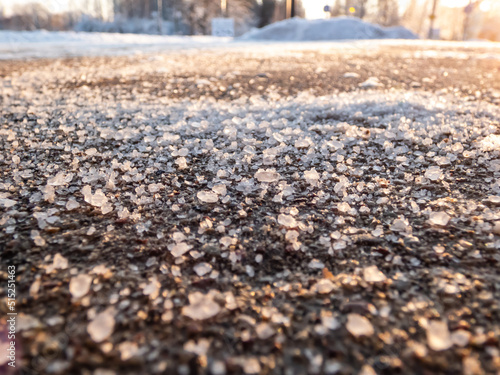 Salt grains on icy sidewalk surface in the winter. Applying salt to keep roads clear and people safe in winter weather from ice or snow. Macro view of salt grains with winter scenery in bacground