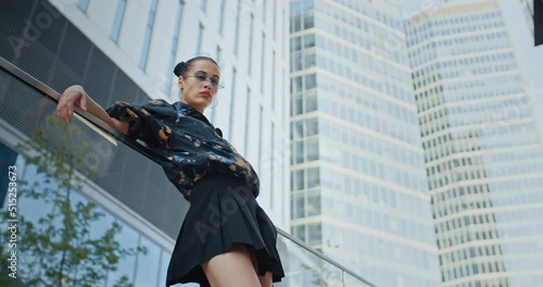 Urban style seductive girl wearing colored jacket short skirt and cool futuristic glasses posing leaning back against glass parapet near high rise skyscrapers. Handsome female fashion model photo