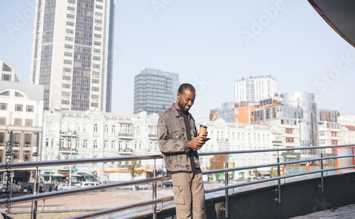 Joyful travel blogger enjoying mobile messaging in social media during leisure for cell networking in city, cheerful African American hipster guy with coffee to go browsing website on smartphone