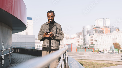 Cheerful dark skinned man in bluetooth earbuds listening positive audio music while chatting with network followers in social media, African American travel blogger enjoying playlist from cellphone