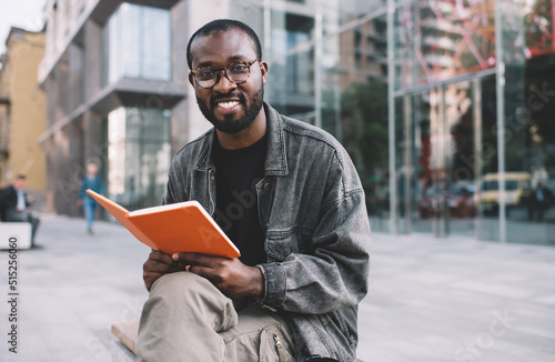 Portrait of carefree male reader in spectacles holding education textbook for learning and posing at city urbanity, cheerful hipster guy in eyewear smiling at camera during leisure with book photo