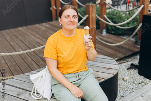 Happy attractive woman siting on bench and eating ice cream on the street. Young smiling girl dressed in yellow t-shirt holding ice cream. Food banner © mdyn