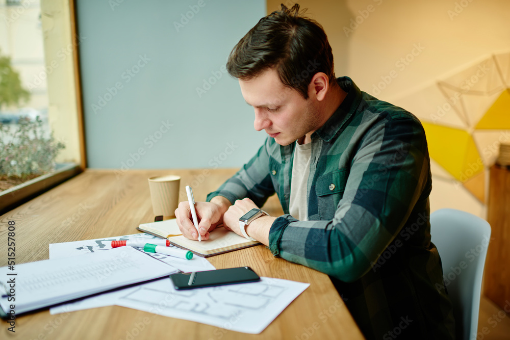 Young male student writing notes in notebook while learning. Man freelancer work on notebook in coffee shop interior, planning schedule Young male student writing notes in notebook while learning. Man freelancer work on notebook in coffee shop interior, planning schedule