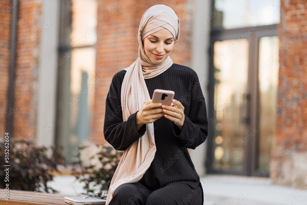 Pretty smiling muslim business woman in hijab sitting on bench and using smart phone during video chat. Happy arabic girl sitting outdoor near her work office and checking email on digital gadget. Pretty smiling muslim business woman in hijab sitting on bench and using smart phone during video chat. Happy arabic girl sitting outdoor near her work office and checking email on digital gadget.