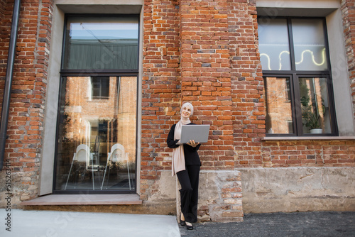 Pretty smiling muslim business woman in hijab standing near office centre and using laptop during video chat. Happy arabic girl standing outdoor near work office and checking email on digital gadget. © sofiko14