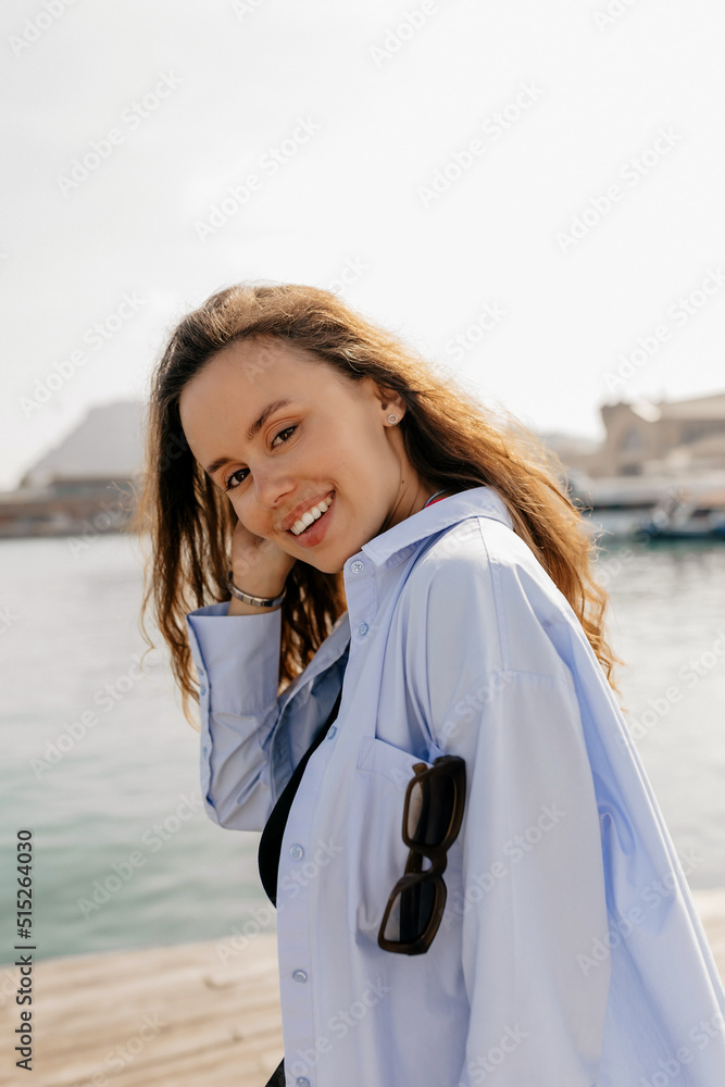 Smiling happy europan girl with long dark hair wearing blue shirt posing at camera on the pier on background of lake in sunlight Smiling happy europan girl with long dark hair wearing blue shirt posing at camera on the pier on background of lake in sunlight