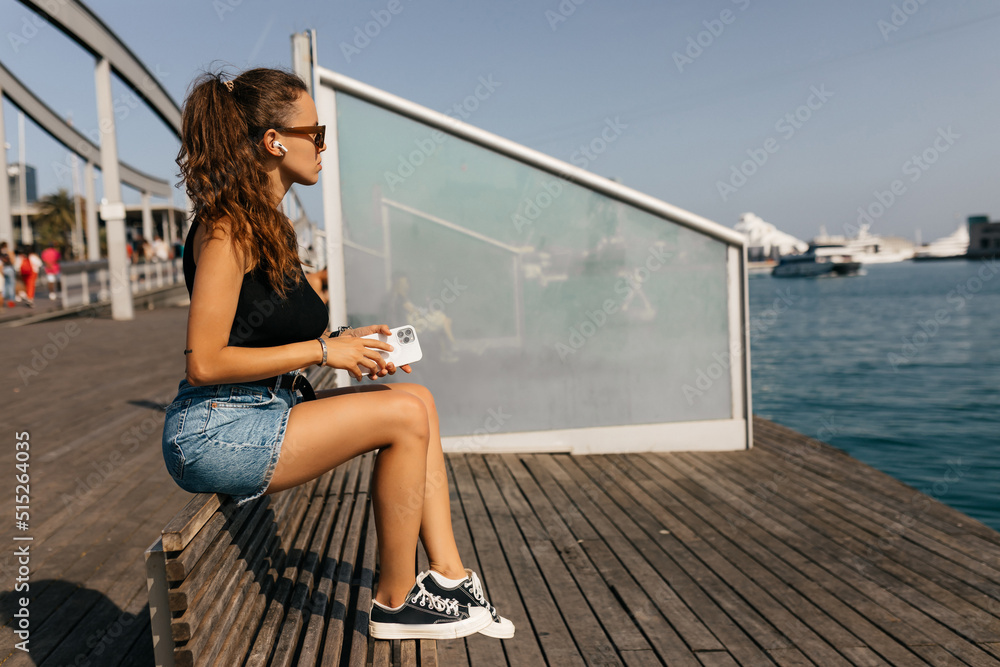 Full-lenght photo of stylish pretty girl in black t-shirt and shorts with long collected hair sitting on the pier in sunlight in warm sunny day