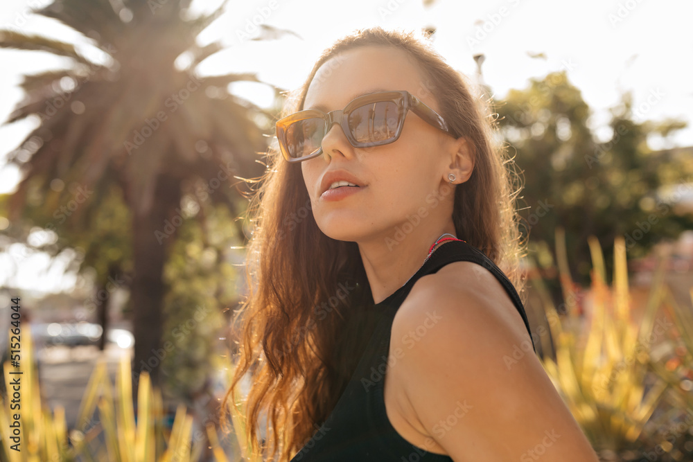 Spectacular lovely woman in sunglasses and black top looking aside and posing in sunshine on background of exotic plants. Optimistic girl with brunette hair smiles and enjoying sun
