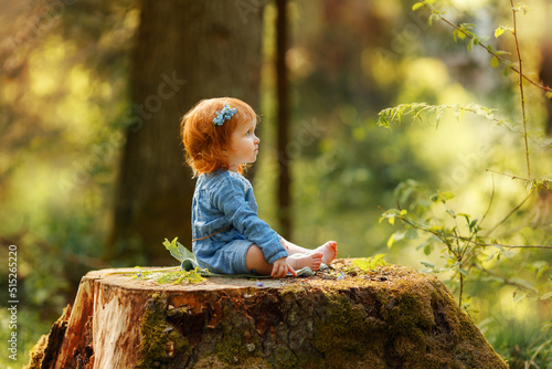 Baby red hair girl in blue dress sitting on a big tree stump in the park  looking at something invisible during sunset. Fantasy concept.