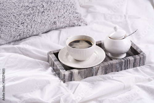 a white porcelain cup with black coffee americano and saucer with sugar in grey marble tray on white bed sheets among fluffy pillows - breakfast in bed
