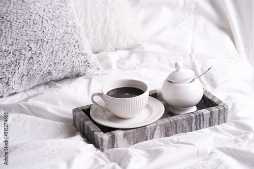 a white porcelain cup with black coffee americano and saucer with sugar in grey marble tray on white bed sheets among fluffy pillows - breakfast in bed