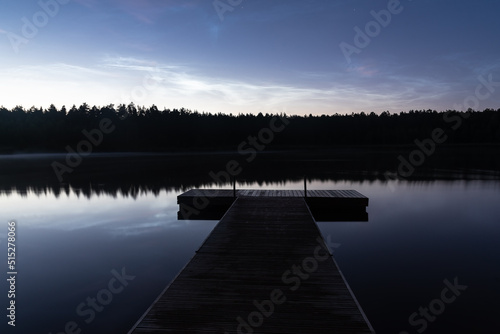Noctilucent clouds over the forest lake in Latvia at night. Wooden pier on foreground.