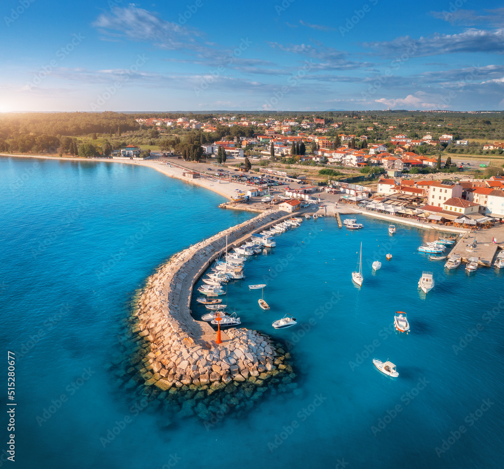 Aerial view of boats and luxure yachts in dock, breakwater, blue sea, old city at sunset in summer. Colorful landscape with sailboats, motorboats, jetty, architecture. Top view of Fazana, Croatia