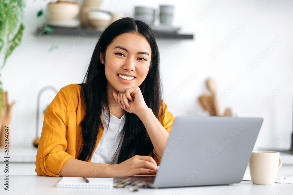 Portrait of lovely positive Asian brunette young woman, creative designer, freelancer or student, sits at the kitchen with a laptop, working or study remotely, looking at the camera, smiling friendly Portrait of lovely positive Asian brunette young woman, creative designer, freelancer or student, sits at the kitchen with a laptop, working or study remotely, looking at the camera, smiling friendly