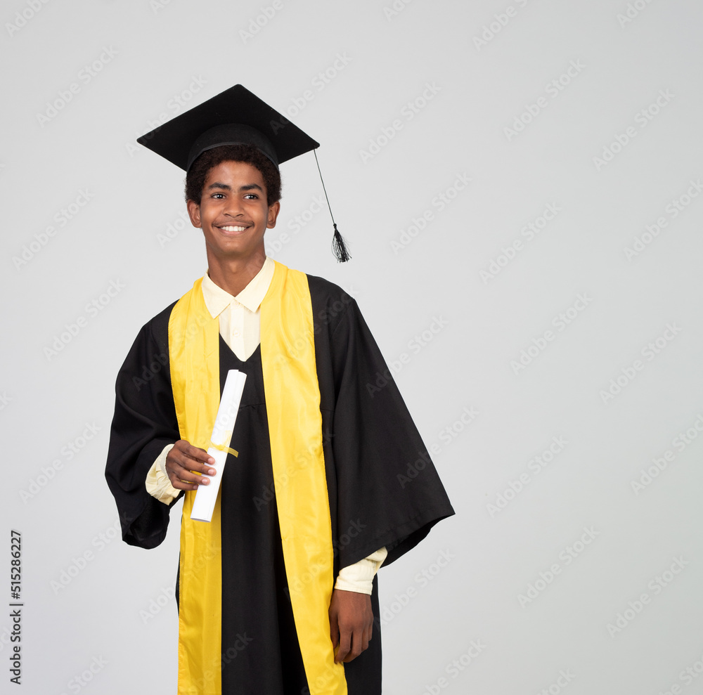 Portrait of graduate teen latin boy student in black graduation gown with  hat, holding diploma - isolated on background. Child back to school and  educational concept. Stock Photo