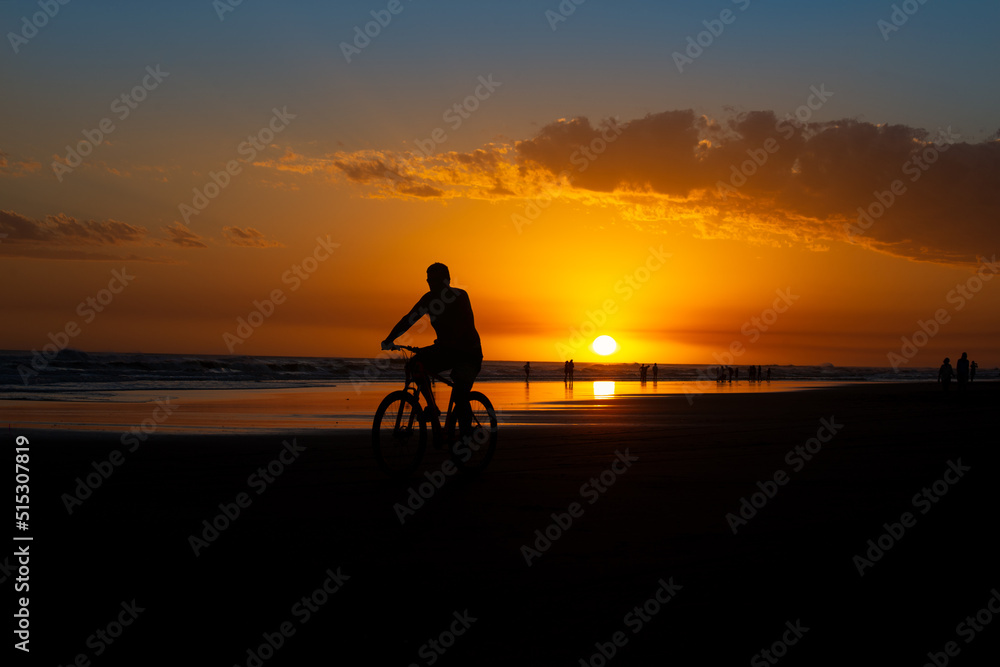 Silhouette of man riding bicycle at sunset along the beach with sun backlighting