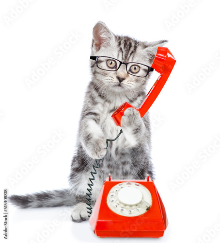 Funny kitten wearing eyeglasses uses a retro phone or telephone. Isolated on white background © Ermolaev Alexandr