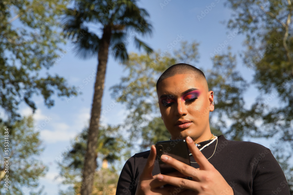 Portrait of non-binary person, young and South American, heavily make up, consulting social networks on his cell phone. Concept queen, lgbtq+, pride, queer.