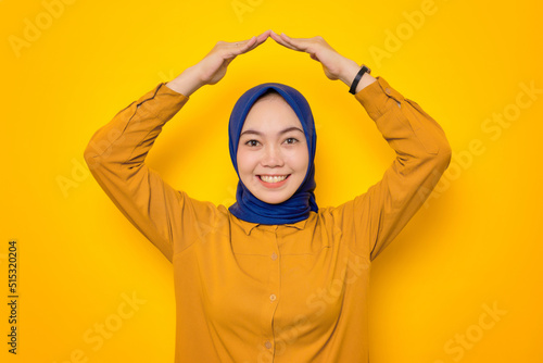 Cheerful young Asian Muslim woman dressed in orange making rooftop home gesture with hands above head isolated on yellow background © Sewupari Studio