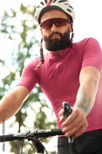 Strong Male cyclist in sportswear, glasses and protective helmet walking with his bike in the forest to take a break after riding. Sky blue and forest in the background. © Georgii
