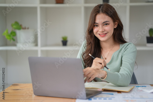 Portrait of young businesswoman using laptop, smiling and looking at camera while sitting at office desk in modern office.
