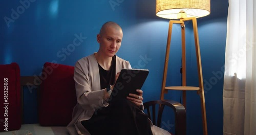 Stylish woman sitting on sofa using ipad and drinking tea at home in evening. Pretty female drawing on tablet screen and reading by lamplight. Working and studying from home office