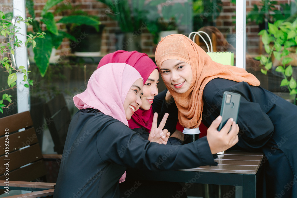 Fototapeta premium Photo of three muslim female students wearing headscarves, sitting on bench in park and taking selfie