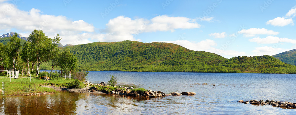 Landscape of a lake with trees, green grass hills. A scenic waterhole surrounded by an empty forest land. Coast of the mountain lake with blue cloudy sky in Nordland near Bodo, Norway