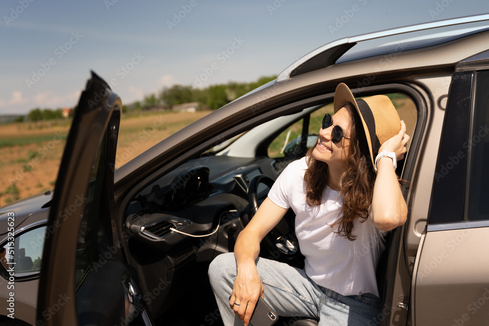 Happy young woman traveler in hat sitting in car on driver's seat in summer in countryside