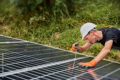 Worker installing photovoltaic solar panel system outdoors. Man engineer placing solar module on metal rails, wearing construction helmets and work gloves, tightening with hex key. © anatoliy_gleb