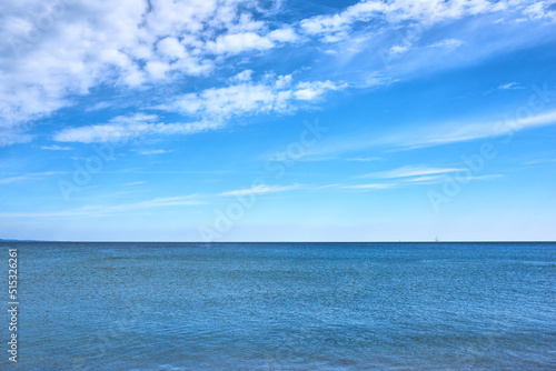 Copy space at sea with a cloudy blue sky background. Calm ocean water across an empty beach with a sailboat cruising in the horizon. Scenic and tranquil landscape view for a peaceful summer holiday