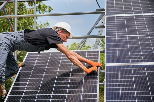 Cropped view of man solar installer placing solar module on metal rails. Male worker wearing safety helmet and work gloves while installing photovoltaic solar panel system outdoors.