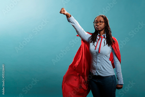 Proud and strong african american justice defender looking ambitious while looking at camera. Powerful and brave young superhero woman wearing hero costume while posing as flying on blue background. photo