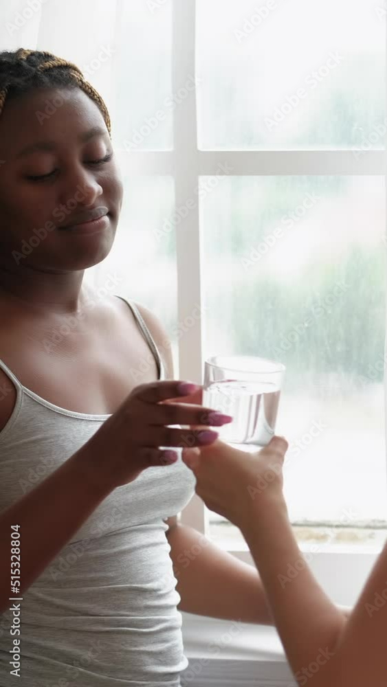 Vertical video. Morning headache. Friend care. Compassion sympathy. Supportive woman bringing glass of water to female roommate suffering from migraine at light white window. Vertical video. Morning headache. Friend care. Compassion sympathy. Supportive woman bringing glass of water to female roommate suffering from migraine at light white window.