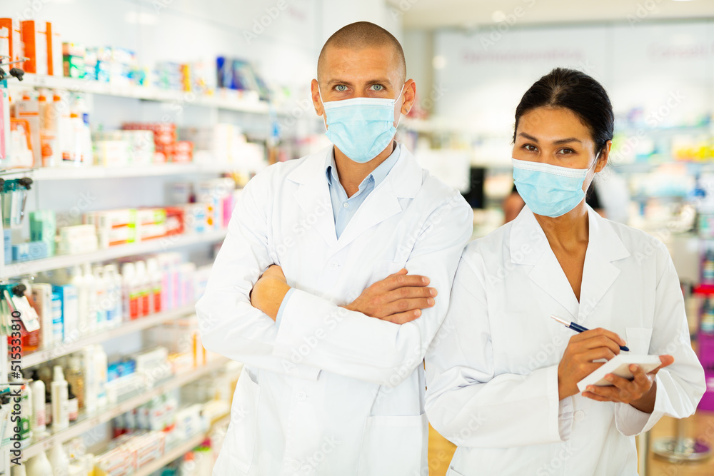Asian woman and European man in doctor's white coats and face masks standing in salesroom of drugstore and looking in camera.