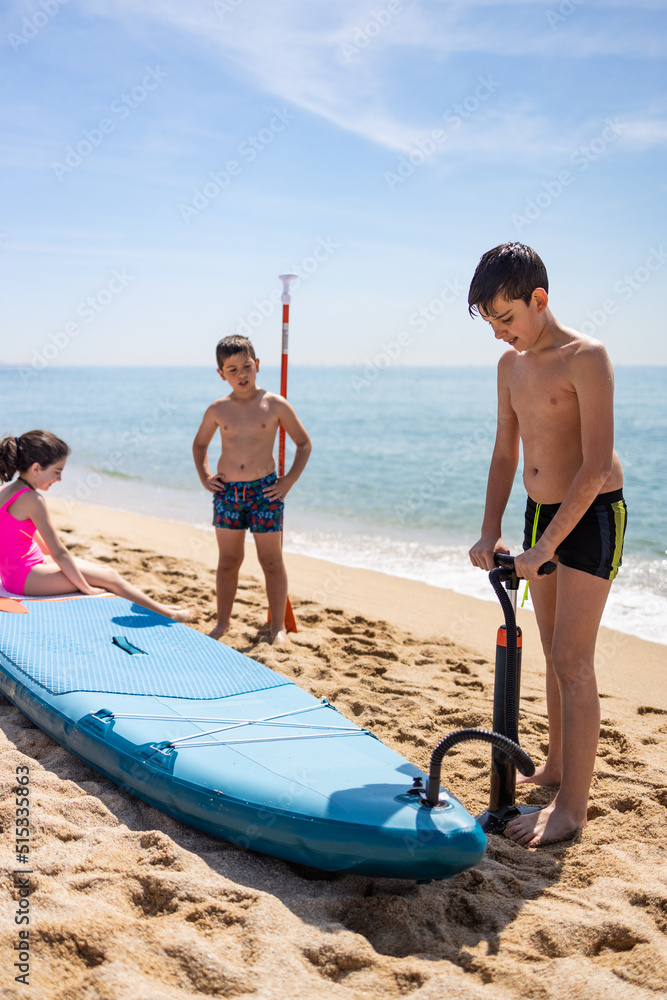 Fototapeta premium Three kids inflating a paddle surf on the beach