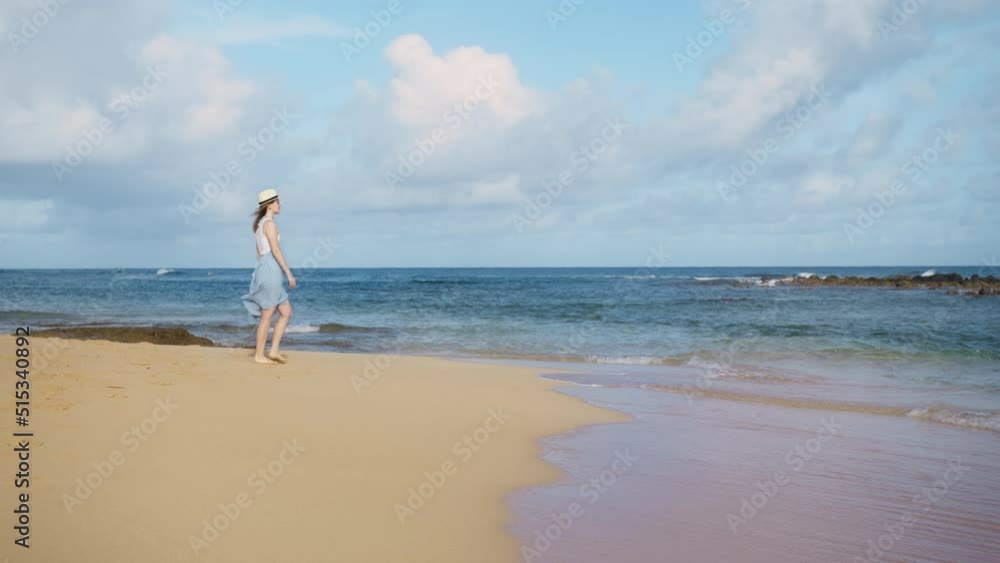 Beautiful slow motion shot of woman in blue flowing beach dress walking barefoot by sandy beach at splashing water waves at calm sunrise. Tourist girl enjoys seascape view on paradise island vacation Beautiful slow motion shot of woman in blue flowing beach dress walking barefoot by sandy beach at splashing water waves at calm sunrise. Tourist girl enjoys seascape view on paradise island vacation