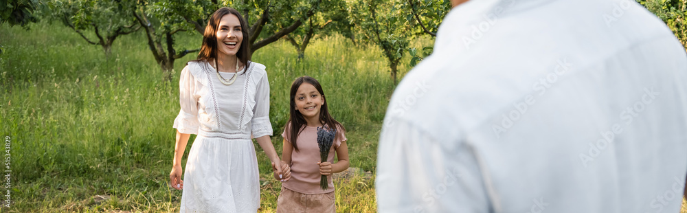 joyful mother and daughter holding hands and smiling near blurred man outdoors, banner.