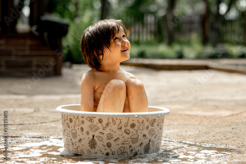 A happy Asian-looking kid with long black hair splashes in a basin in the yard of a house on a hot summer day. The concept of a happy childhood