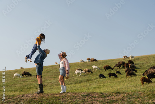 side view of farmer raising up daughter near wife and herd grazing in green pasture. © LIGHTFIELD STUDIOS
