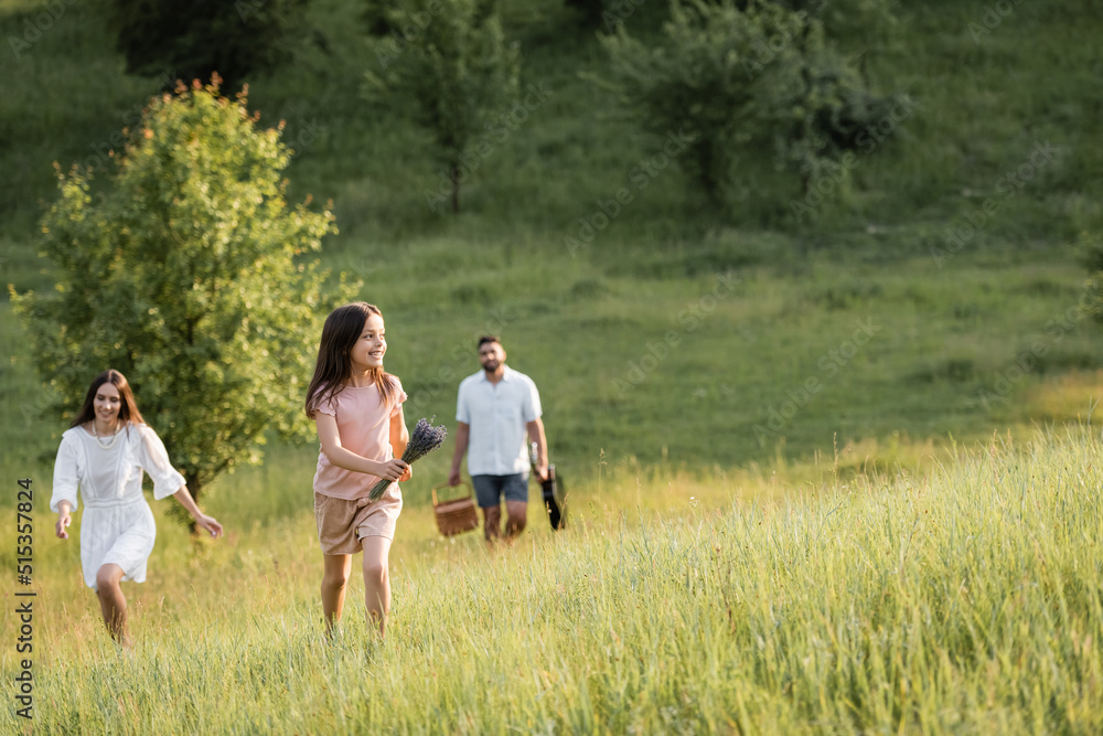 happy child with lavender bouquet running in meadow near parents on blurred background.