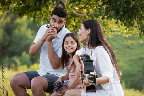 happy couple playing guitar and harmonica near cheerful daughter outdoors.