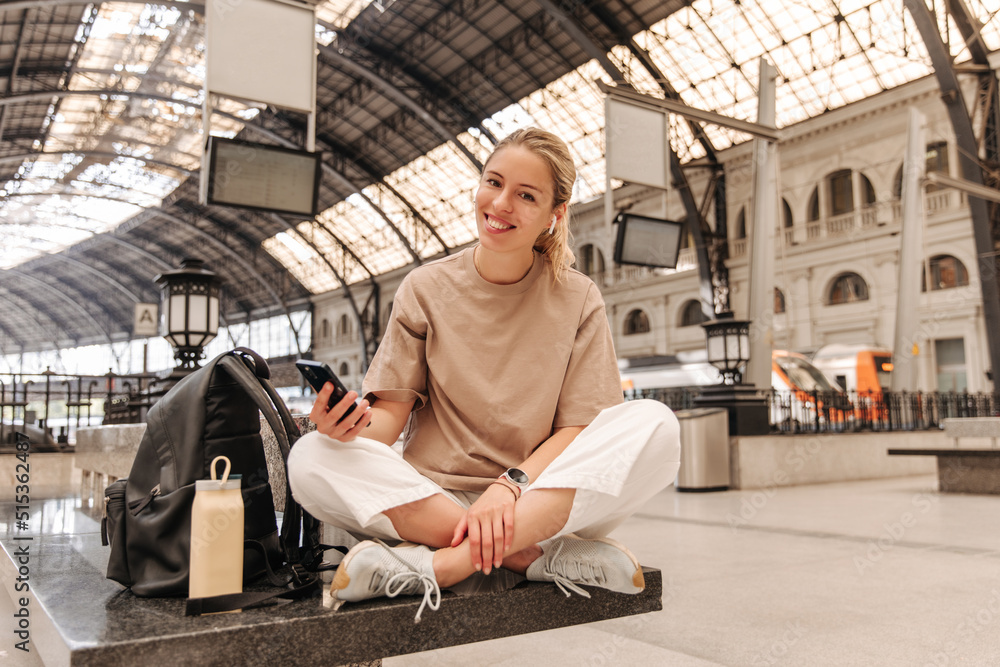 Full view of pretty caucasian woman holding phone and sitting on the bench . Blonde cute lady look with smile at camera with backpack 