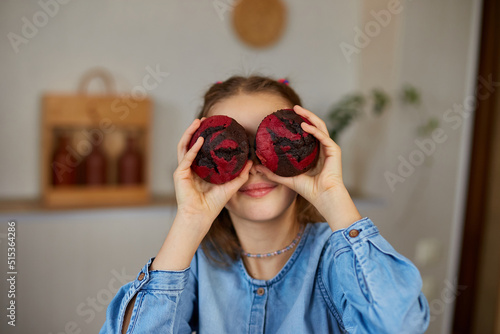 Happy emotional little girl holding cupcakes in hands covering eyes at home