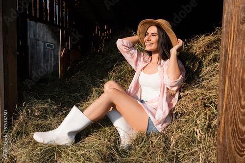 Foto smiling woman sitting on haystack in rubber boots and adjusting straw hat