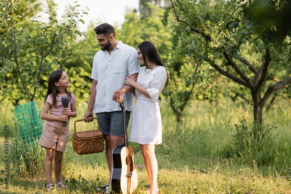 happy couple with wicker basket and acoustic guitar looking at daughter in countryside garden. happy couple with wicker basket and acoustic guitar looking at daughter in countryside garden.