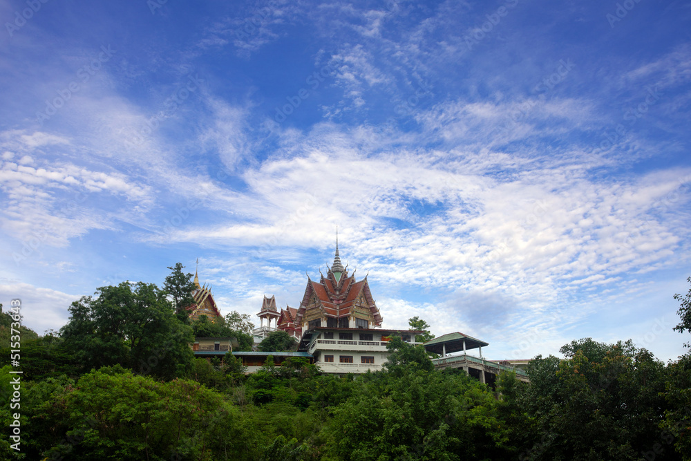 Thai temple is located on hillside surrounded by nature of forest and bright blue sky.