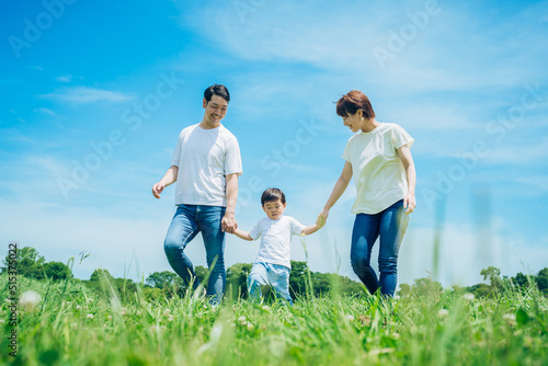 手を繋いで晴天の緑地を歩く親子3人 © maru54