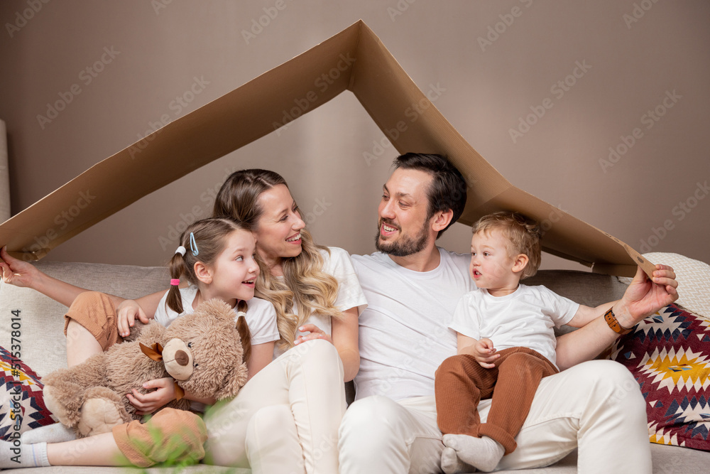 Family complete parents with two children sitting on floor relaxing after moving to new apartment holding piece of cardboard left over from boxes as roof over their heads smiling to camera each other.