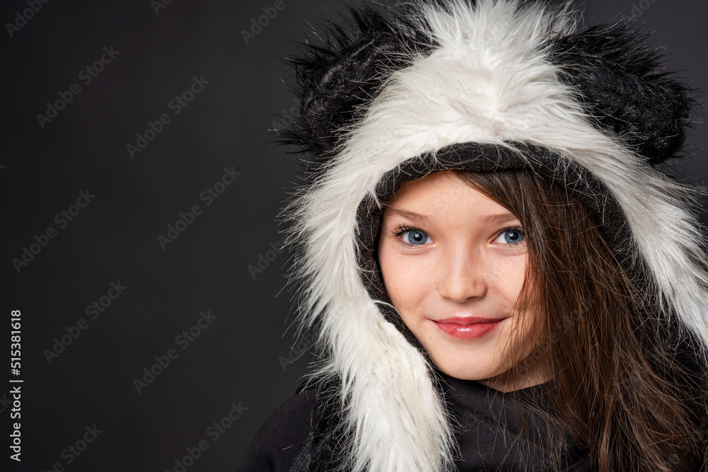 Portrait of a cheerful cute teenage girl with long black hair, dressed in a panda fur hat, posing on a black studio background. A comical image. Advertisement. original children's clothing. Close-up Portrait of a cheerful cute teenage girl with long black hair, dressed in a panda fur hat, posing on a black studio background. A comical image. Advertisement. original children's clothing. Close-up